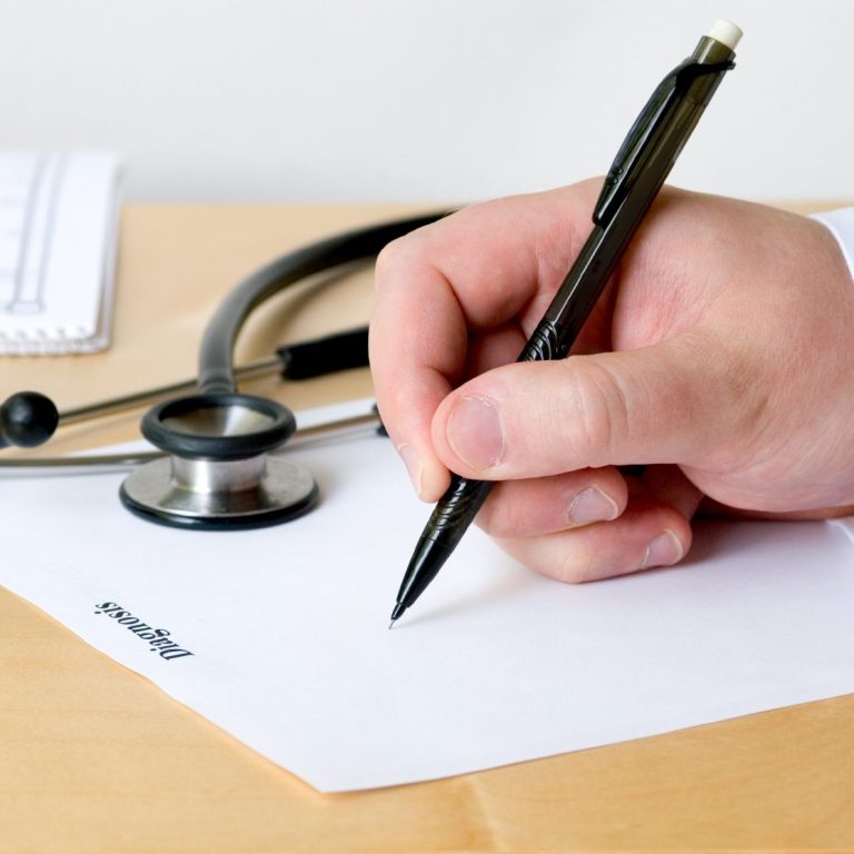 Hand mit einem Stift, die auf ein leeres Papier neben einem Stethoskop schreibt.